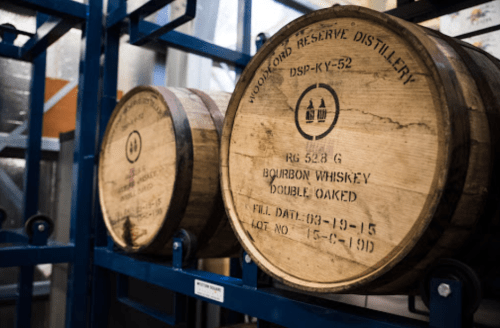 Two wooden barrels labeled "Woodford Reserve Distillery" stacked on a rack, containing bourbon whiskey.