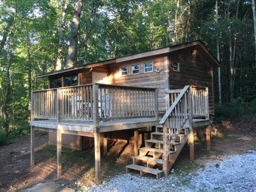 A wooden cabin on stilts surrounded by trees, featuring a porch and steps leading up to the entrance.