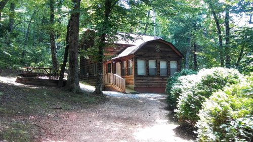 A rustic wooden cabin surrounded by trees, with a gravel path leading to the entrance.