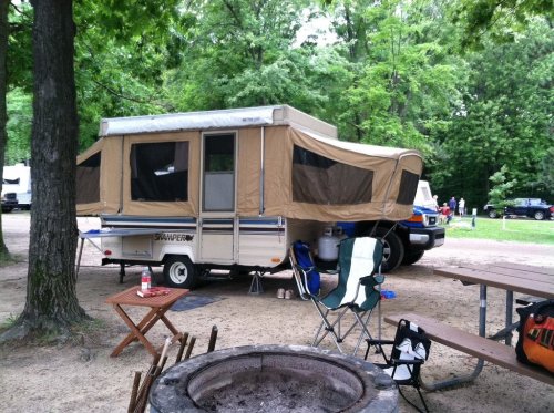 A pop-up camper parked in a wooded campsite, with a fire pit and picnic table nearby.