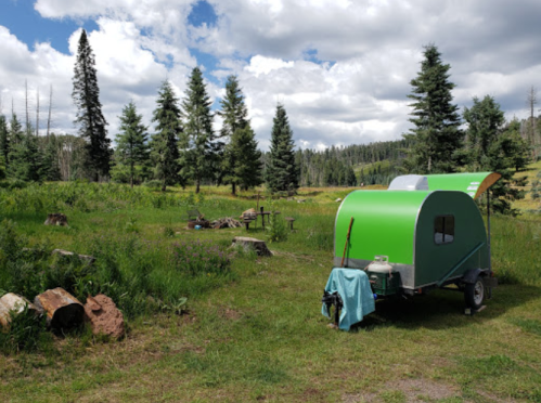 A green camper trailer parked in a grassy area surrounded by tall trees and a blue sky with fluffy clouds.