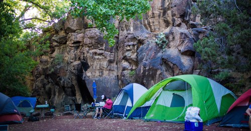 A campsite with green and blue tents set against a rocky backdrop, surrounded by trees and outdoor cooking equipment.