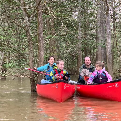 A family kayaking in red canoes on a calm, wooded lake, surrounded by trees and greenery.