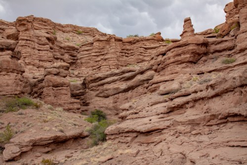 Rocky desert landscape with layered formations and sparse vegetation under a cloudy sky.