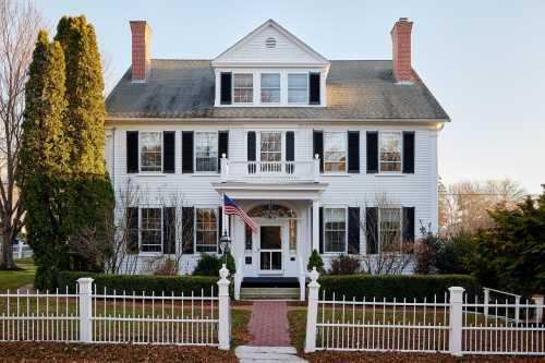 A large white colonial-style house with black shutters, a front porch, and an American flag, surrounded by a white picket fence.