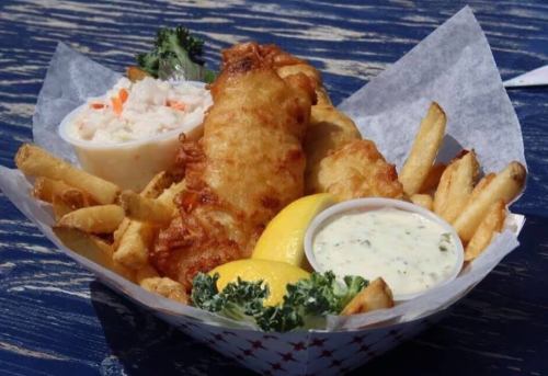 A basket of crispy fish and fries, served with coleslaw, lemon wedges, and tartar sauce on a blue wooden table.