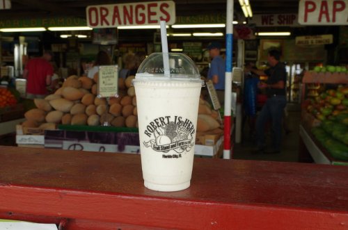 A milkshake in a cup with a logo, placed on a counter in a vibrant fruit market. Oranges and produce in the background.