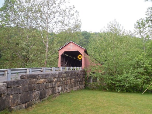A red covered bridge surrounded by greenery, with a stone wall and a railroad crossing sign nearby.