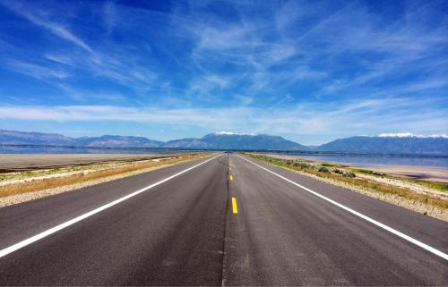 A straight road stretches into the distance, flanked by mountains and a clear blue sky with wispy clouds.
