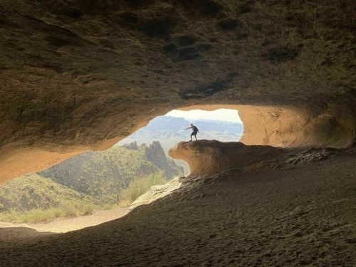 A person stands on a rock ledge inside a cave, framed by a large opening with a scenic view of mountains outside.