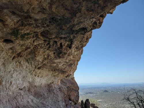 A rocky cliff overhang with a clear blue sky and a distant view of a valley below.