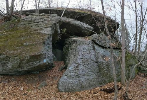 A large rock formation with a cave-like opening, surrounded by trees and fallen leaves in a wooded area.