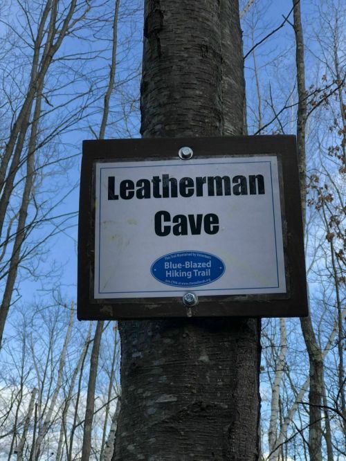Sign on a tree marking "Leatherman Cave" along a blue-blazed hiking trail, surrounded by bare trees and a clear sky.
