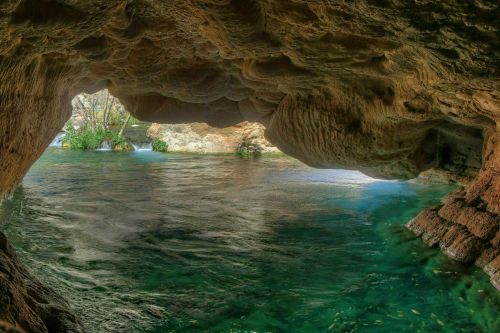 A serene cave opening reveals clear turquoise water and lush greenery beyond, creating a tranquil natural scene.