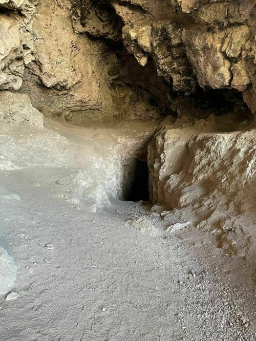A rocky cave interior with a dark entrance and sandy floor, illuminated by natural light.