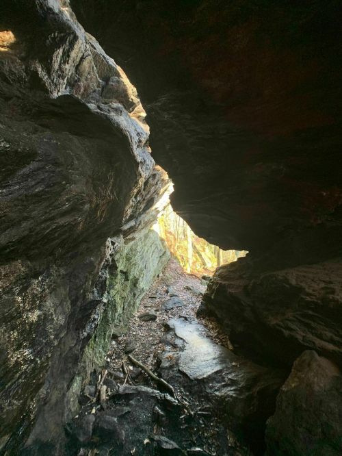 A narrow cave opening with sunlight streaming through, revealing a rocky path and lush greenery outside.