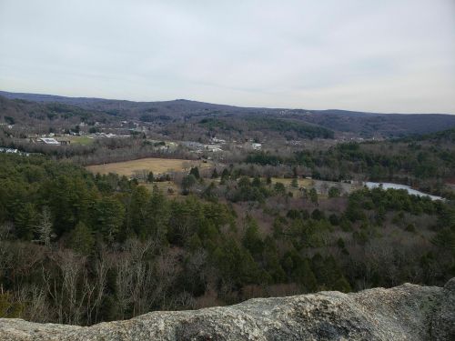 A panoramic view of a valley with trees, fields, and a small lake, surrounded by rolling hills under a cloudy sky.