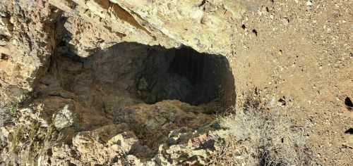 A deep, rocky cave entrance surrounded by dry earth and sparse vegetation.