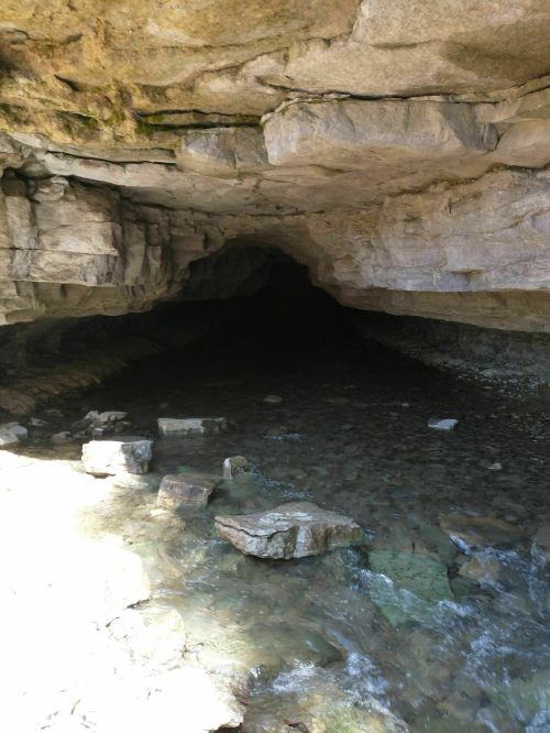 A rocky cave entrance with a small stream flowing in front, surrounded by natural stone formations.