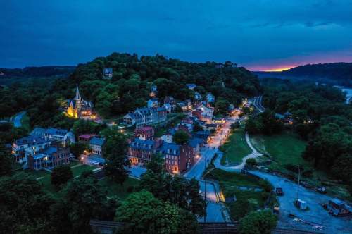 Aerial view of a quaint town at dusk, with illuminated buildings and a church on a hillside surrounded by trees.
