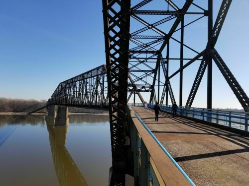 A metal bridge spans a calm river, with a person walking along the pathway under a clear blue sky.