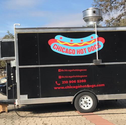 A food truck with a black exterior featuring "Chicago Hot Dogs" and colorful hot dog graphics. Contact info displayed.