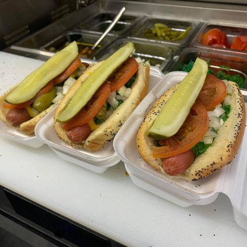 Three hot dogs in buns topped with tomatoes, pickles, onions, and green relish, served in takeout containers.