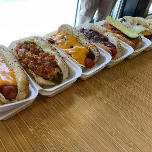 A row of hot dogs in buns, topped with cheese, chili, and pickles, served in white containers on a bamboo surface.