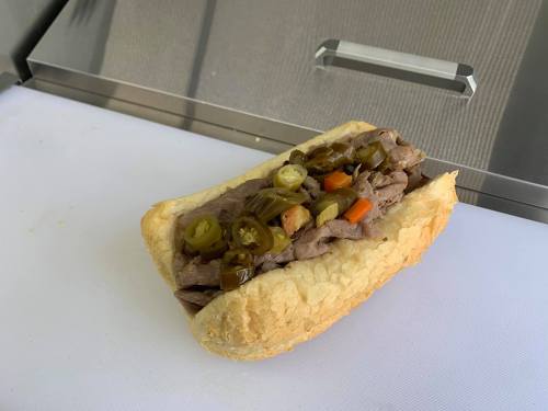 A sandwich filled with sliced beef, jalapeños, and vegetables on a crusty roll, resting on a white cutting board.