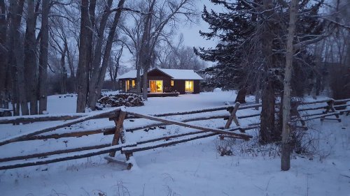 A cozy cabin with glowing windows surrounded by snow-covered trees and a wooden fence in a winter landscape.