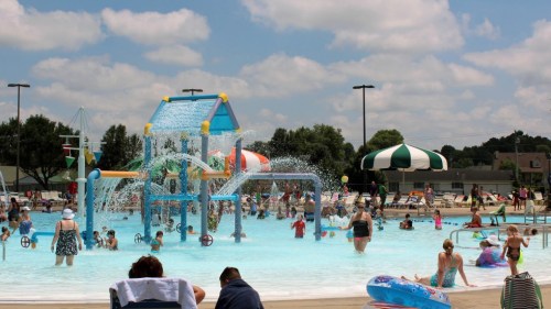 A busy outdoor pool scene with families enjoying water play, a splash pad, and colorful umbrellas under a blue sky.