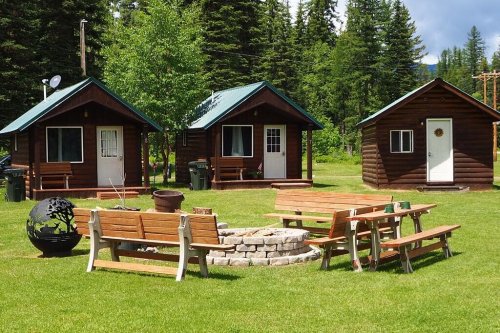Three rustic cabins surrounded by green grass, picnic tables, and a stone fire pit in a wooded area.