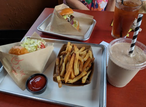 Two burgers in paper wrappers, a side of fries with ketchup, and a drink with a straw on a red table.
