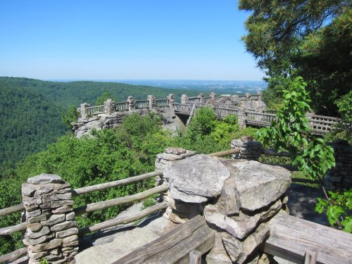 Scenic view from a stone overlook, surrounded by lush greenery and mountains under a clear blue sky.