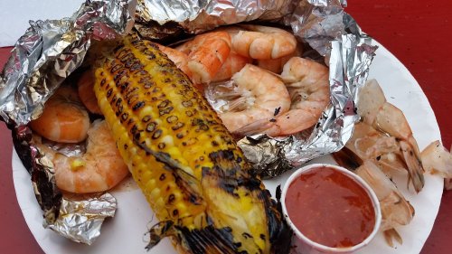 A plate with grilled corn on the cob, shrimp, and a small cup of dipping sauce, all wrapped in foil.