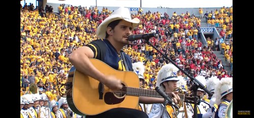 A musician in a cowboy hat plays guitar and sings on stage, with a large crowd in the background.