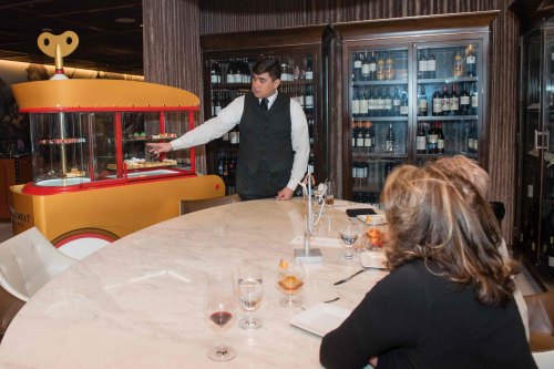 A waiter presents desserts from a colorful cart to guests seated at a marble table in an elegant restaurant.