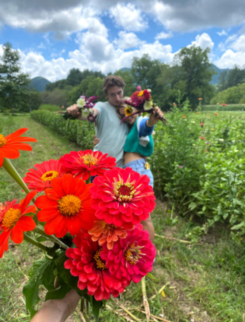 A person holds vibrant red and pink flowers in the foreground, while two friends pose playfully in a flower field behind them.