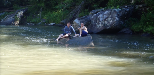 Two people sit on a rock in a river, surrounded by greenery, enjoying a sunny day by the water.