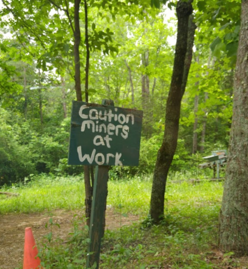 A wooden sign reading "Caution miners at work" stands in a green, wooded area.