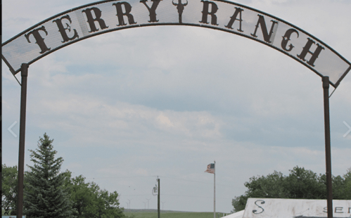 Sign for Terry Ranch with a cloudy sky and an American flag in the background. Green trees are visible nearby.
