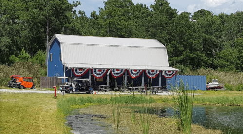 A blue barn-style building decorated with red, white, and blue bunting, near a pond and surrounded by greenery.