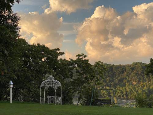 A white gazebo and swing set on a grassy area, with lush trees and a scenic view of clouds and hills in the background.