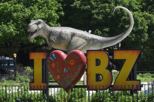 A large dinosaur statue stands behind a colorful "I ❤️ BZ" sign in a park setting with green trees.