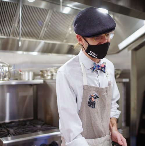 A chef in a mask and bow tie prepares food in a modern kitchen, wearing an apron with a logo.