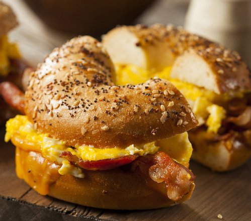 A toasted everything bagel sandwich filled with scrambled eggs, cheese, and crispy bacon on a wooden surface.
