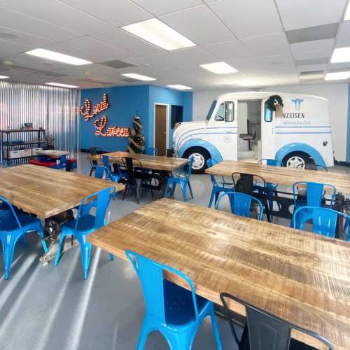 A bright, modern dining area with wooden tables, blue chairs, and a vintage food truck in the corner.