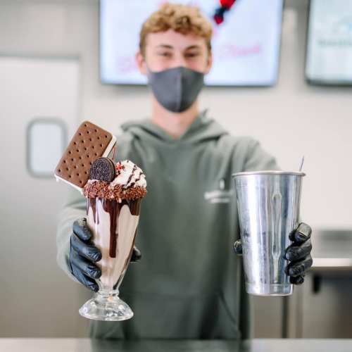 A person in a mask holds a tall ice cream sundae topped with a cookie and a metal cup in a dessert shop.