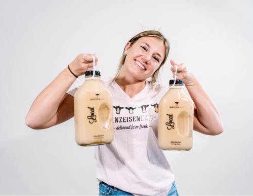 A smiling woman holds two large bottles of milk, wearing a t-shirt with the logo of a dairy farm.