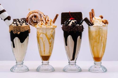 Four tall glasses of decadent milkshakes topped with various sweets, including cookies, chocolate, and whipped cream.